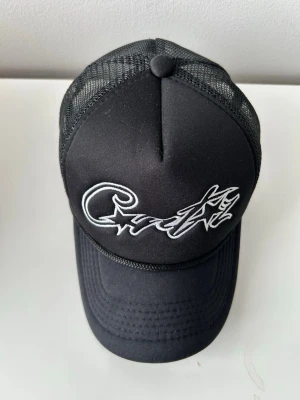 Corteiz keps - Snygg svart truckerkeps med vit broderad text och stjärna framtill. Kepsen har mesh-bakstycke för extra ventilation och justerbar snapback. Perfekt för en streetwear-look och passar till de flesta outfits.
