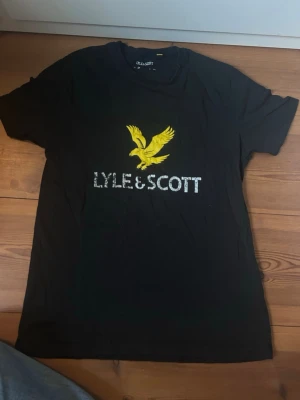 Svart Lyle & Scott t-shirt med tryck - Snygg svart t-shirt från Lyle & Scott med stort gult örntryck och vit logga på bröstet. Klassisk passform och rund halsringning, perfekt till jeans eller shorts. Jättebra skick! Skriv om du har frågor.