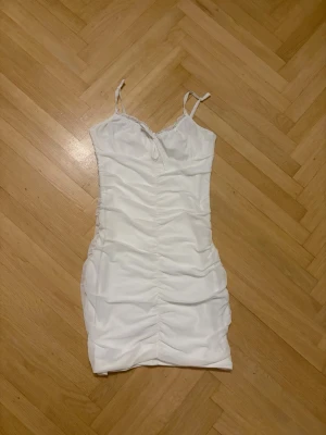 Vit kort klänning med rynkade detaljer - Säljer en vit kort klänning med smala axelband och rynkade detaljer längs hela klänningen. Klänningen har en figurnära passform och är tillverkad i ett mjukt material som känns bekvämt mot huden. Perfekt för dig som gillar en enkel men snygg stil. Fin på sommaren, våren och skolavslutningen. 