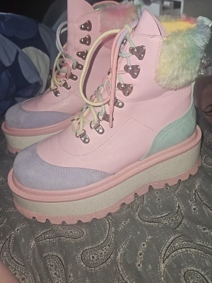 Pastellrosa platå-kängor med fluff - Säljer ett par unika pastellrosa kängor med tjock platåsula och regnbågsfärgat fluff runt skaftet. Skorna har metallhål för snörning, detaljer i ljuslila och mintgrönt samt en rejäl sula i rosa och beige. Perfekta för dig som vill sticka ut med färg och cool stil. Ny pris 2000kr