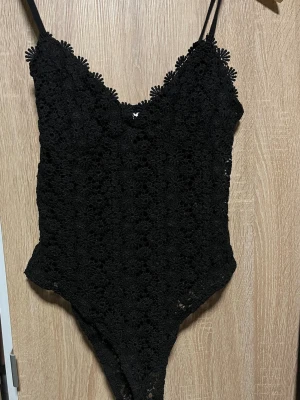 Svart body med blommönster - Säljer en svart body med tunna axelband och vadderade kupor. Bodyn är täckt av ett detaljerat blommigt spetsmönster och har en feminin, figurnära passform. Perfekt för dig som gillar en elegant och trendig look.