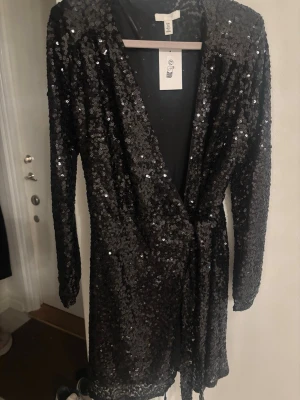 Svart paljettklänning från H&M - Säljer en svart omlottklänning från H&M täckt med glittriga paljetter. Klänningen har lång ärm, v-ringning och ett bälte i midjan för snygg passform. Perfekt för dig som vill sticka ut med en riktigt festlig look.