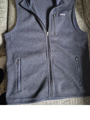 Blå dunväst från Patagonia - Snygg blå dunväst från Patagonia med hel dragkedja framtill, två sidofickor med dragkedja och en bröstficka. Västen har en hög krage och är tillverkad i ett mjukt, strukturerat material. Perfekt för lager-på-lager och chill stil.