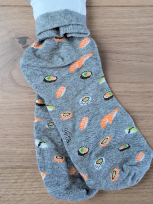 Gråa strumpor med sushimönster - Gråa strumpor med färgglatt sushimönster i orange, vitt och grönt. Perfekta för dig som vill ha något lekfullt och unikt på fötterna. Strumporna är låga och tillverkade i mjuk bomullsmix för skön komfort hela dagen.