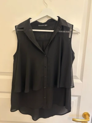 Svart ärmlös blus från Pull&Bear - Snygg svart ärmlös blus från Pull&Bear i tunn, transparent chiffong. Blusen har knappar framtill, klassisk krage och ett lager över bröstet som ger en cool, flowy look. Perfekt att styla med jeans eller kjol för en trendig outfit.