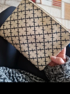 Beige kuvertväska från By Malene Birger - Snygg beige kuvertväska från By Malene Birger med klassiskt svart blommönster och guldfärgad dragkedja. Väskan är i tåligt konstläder och har logga framtill. Perfekt att bära till allt och ger en trendig touch till din outfit.