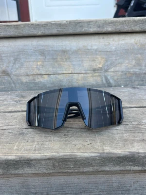 Svarta sportiga solglasögon - Säljer ett par svarta sportiga solglasögon med bred, rektangulär lins och futuristisk design. Glasögonen har en skarp, kantig form och passar perfekt för dig som gillar en modern och cool stil. Ramen är i svart plast och linsen är mörktonad.