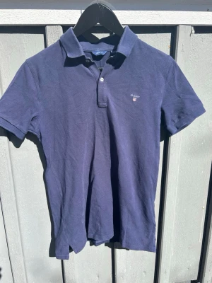 Marinblå pikétröja från Gant - Klassisk marinblå pikétröja från Gant med korta ärmar och liten logga broderad på bröstet. Tröjan har krage och två knappar vid halsen. Tillverkad i mjuk bomull som känns skön mot huden. Perfekt till jeans eller shorts.