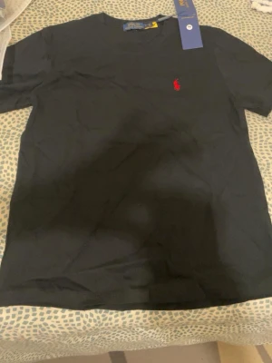 Svart t-shirt från Polo Ralph Lauren - Klassisk svart t-shirt från Polo Ralph Lauren med den ikoniska röda logotypen broderad på bröstet. T-shirten har rund hals och korta ärmar. Tillverkad i mjuk bomull för en skön känsla och enkel stil.