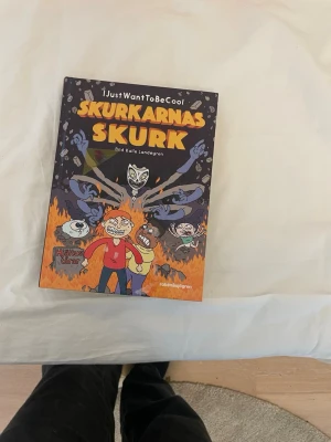 Skurkarnas Skurk av IJustWantToBeCool - En färgstark bok med tecknade illustrationer av Kalle Landegren. Omslaget är mörkt med orange och gula eldflammor, och boken har en cool, humoristisk ton. Perfekt för dig som gillar äventyr och skurkar i serieformat.