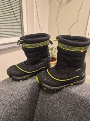 Svarta vinterstövlar från Viking 28 - Säljer ett par svarta vinterstövlar från Viking med gröna detaljer och dragsko upptill. Stövlarna har grov sula för bra grepp och är tillverkade i slitstarkt material som tål snö och slask. Perfekta för kalla dagar och håller fötterna torra och varma.