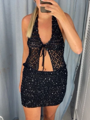 Svart glittrigt set - Säljer ett svart glittrigt set med halterneck topp och öppen front som knyts med snöre. Settet har ett virkat utseende och är figurnära med en snygg, festlig vibe. Perfekt för sommarens festligheter!