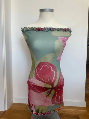 Blommig offshoulder kortklänning - Trendig kortklänning med offshoulder-design och stora blommiga mönster i rosa och rött på en ljusgrön bakgrund. Klänningen har volangdetaljer vid halsringning och nederkant, och är figurnära med stretchigt material.