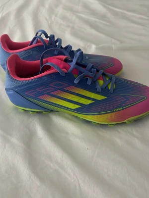 Adidas f50 fotbollsskor - Säljer ett par Adidas fotbollsskor i blått, rosa och neongult. Skorna har klassiska tre ränder på sidan, snörning och en sula med dobbar i neongult för bra grepp på planen. Yttermaterialet är syntetiskt och designen är sportig och modern. Dubbarna är Ag dubbar (konstgräs) men funkar också på vanligt naturgräs. Skorna är använda en gång och är i storlek 40. 
