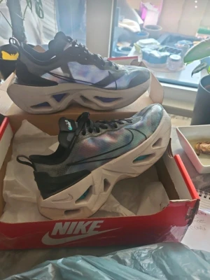 Nike sneakers med chunky sula - Säljer ett par Nike sneakers med futuristisk chunky sula och svart, blå och lila mesh-överdel. Skorna har svarta detaljer, snörning och en unik design med genomskinliga partier i sulan. Perfekta för dig som vill sticka ut med din stil.