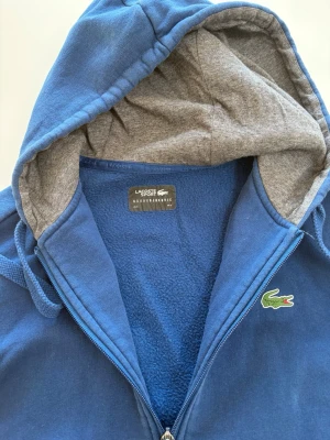 Blå zip hoodie från Lacoste - Blå hoodie från Lacoste med dragkedja och grått innerfoder i huvan. Klassisk krokodillogga på bröstet och två fickor framtill. Tillverkad i mjukt bomullsmaterial. 