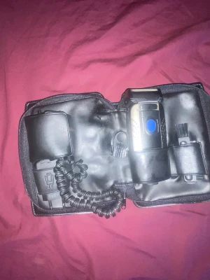 Braun rakapparat i svart fodral - Säljer en elektrisk rakapparat från Braun i ett svart fodral. Setet innehåller rakapparat, laddare och rengöringsborste. Rakapparaten har silverfärgad kropp med blå knapp och detaljer. Perfekt för grooming och enkel att ta med på resan.