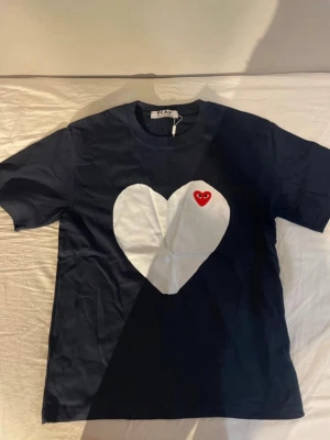 Marinblå Comme des Garçons Play t-shirt - Snygg marinblå t-shirt från Comme des Garçons Play med stort vitt hjärta och det klassiska röda hjärtloggan på bröstet. T-shirten har rund hals och korta ärmar, perfekt för en clean och trendig look. Ser använd ut på bilden, men den är helt oanvänd. Priset är ej hugget i sten. 