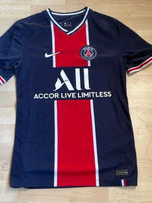PSG matchtröja Nike Dri-FIT blå/röd - Snygg PSG matchtröja från Nike i Dri-FIT material. Tröjan är mörkblå med bred röd mittremsa, vita detaljer och klubbmärke på bröstet. V-ringad krage med vita och röda ränder. Perfekt för fotbollsträning eller att bära som supporter.