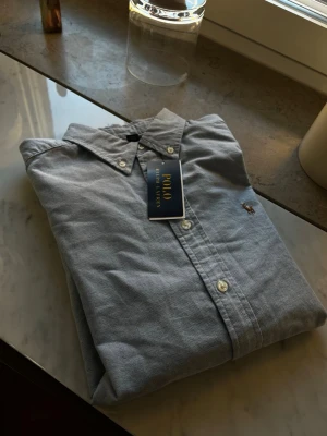 Blå skjorta från Polo Ralph Lauren - Helt oanvänd klassisk blå skjorta från Polo Ralph Lauren med button-down krage och broderad logga på bröstet. Skjortan har vita knappar och är tillverkad i bomull för en bekväm känsla. Perfekt till jeans eller chinos för en clean look.