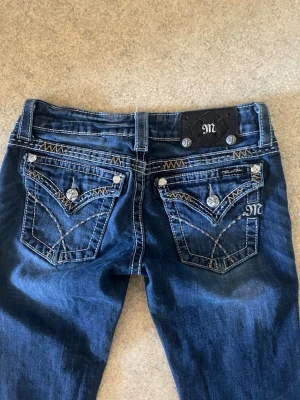 Mörkblå Miss Me bootcut jeans - Snygga mörkblå jeans från Miss Me med bootcut passform. Jeansen har dekorativa bakfickor med broderier och blingknappar samt kontrastsömmar. Klassisk femficksmodell i slitstark denim med detaljer som sticker ut.