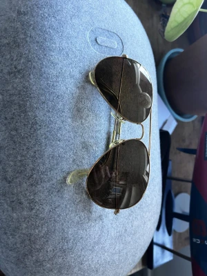 Ray-Ban Aviator solglasögon guld - Klassiska Ray-Ban Aviator solglasögon med guldfärgad metallbåge och bruna linser. Säljs med original Ray-Ban fodral i mörkbrunt skinn. Perfekt accessoar för en cool och tidlös stil.