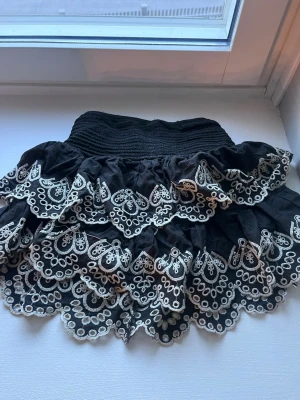 Svart volangkjol med spets från Lindex - Supersöt svart kjol från Lindex med flera lager volanger och vit broderad spetskant. Kjolen har bred resår i midjan för skön passform och ger en riktigt lekfull och trendig look. Perfekt för dig som gillar detaljer och vill sticka ut lite extra.