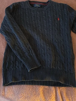 Mörkblå kabelstickad tröskel Ralph lauren - Kabelstickad mörkblå tröja från Polo Ralph Lauren med rund halsringning och röd broderad logga på bröstet. Tröjan har ribbade muddar vid ärmslut och nederkant. Perfekt för lager på lager och klassisk stil.