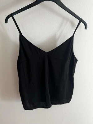 Svart linne med smala axelband Lindex - Snyggt svart linne från Lindex med smala axelband och v-ringad hals. Perfekt att styla till jeans eller kjol. Tillverkat i ett mjukt och lätt material som känns skönt mot huden.