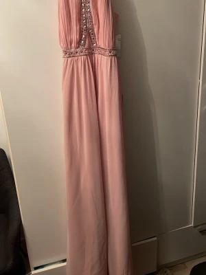 Ljusrosa långklänning med pärldetaljer - Elegant ljusrosa långklänning med vackra pärldetaljer och paljetter vid midjan och överdelen. Klänningen har ett plisserat liv och är axelbandslös, vilket ger en stilren och festlig look. Perfekt för bal eller speciella tillfällen.