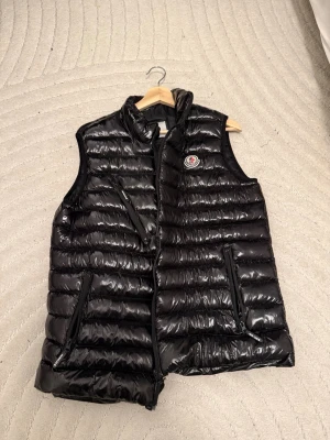 Svart dunväst från Moncler - Snygg svart dunväst från Moncler med glansig finish och klassisk logga på bröstet. Västen har två dragkedjeförsedda fickor och hög krage. Perfekt för lager-på-lager och ger en sportig vibe till din outfit.