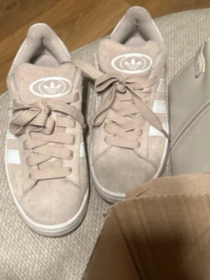 Beige Adidas sneakers i mocka - Snygga beige sneakers från Adidas i mjuk mocka med klassiska vita ränder på sidorna och Adidas-logga på plösen. Skorna har beige snörning och vit sula. Perfekta för dig som gillar stilrena och bekväma sneakers med retrovibbar. Har haft på mig de max 2 gånger 