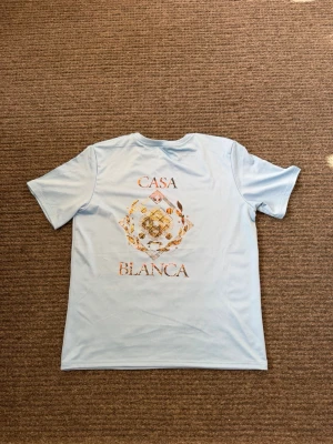 Ljusblå Casa Blanca t-shirt - Snygg ljusblå t-shirt från Casa Blanca med ett stort, detaljerat tryck i guld och orange på ryggen. Trycket har ett grafiskt mönster och texten 'CASA BLANCA'. Perfekt för dig som vill sticka ut med en unik tee.