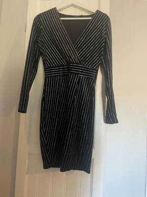 Svart omlottklänning med silverränder - Snygg svart kortklänning med omlott och v-ringning, med tunna silverfärgade ränder. Klänningen har lång ärm och markerad midja. Använd max 3 gånger.