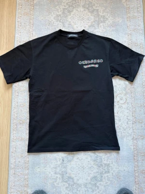 Svart Chrome Hearts t-shirt - Säljer en svart t-shirt från Chrome Hearts med färgglada broderade detaljer fram och stort korsmönster på ryggen. Klassisk passform och rund halsringning. Perfekt för dig som gillar streetwear och vill sticka ut lite extra.
