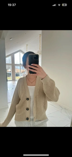 Beige stickad kofta med stora knappar - Säljer en beige stickad kofta med v-ringning och stora svarta knappar. Koftan har ribbade muddar och en avslappnad passform. Perfekt att slänga över ett linne eller t-shirt för en chill look.