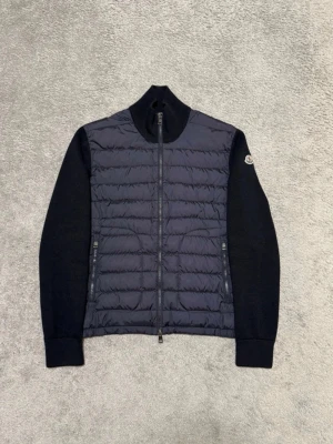 Moncler cardigan - Snygg mörkblå kofta från Moncler med quiltad, vadderad front och stickade ärmar. Skicket är 10/10 den är inte använd. Dragkedja framtill och två sidofickor med dragkedja. Moncler-logga på vänster ärm. Perfekt för lager-på-lager.