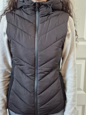 Svart dunväst med huva - Snygg svart dunväst med dragkedja framtill och stor, vadderad huva. Västen har ett quiltat mönster i chevron-stil och sidopaneler i stretchigt material för extra komfort. Perfekt för lager-på-lager och håller dig varm under kyliga dagar.