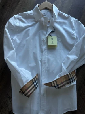 Vit skjorta från Burberry med rutiga detaljer - Klassisk vit skjorta från Burberry med ikoniska beige-rutiga detaljer på manschetterna. Skjortan har lång ärm, knappar framtill och diskret broderad logga på bröstet. Perfekt för dig som gillar stilrena plagg med exklusiva detaljer.