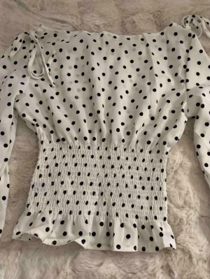 Vit prickig topp med smock - Trendig populär vit topp med svarta prickar och smockad midja som ger en snygg siluett. Långa ärmar och volangkant nedtill för en feminin touch. Tillverkad i ett lätt och mjukt material som känns skönt mot huden. Perfekt att styla med jeans eller kjol.