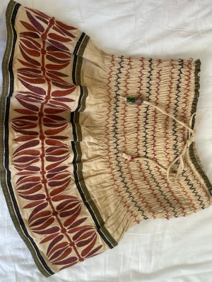 Beige kortkjol med rött mönster - Unik beige kortkjol med röda och svarta broderade detaljer och elastisk smock i midjan. Kjolen har ett dekorativt snöre med pärlor och ett grafiskt mönster längs fållen. Obs!! Den hade tidigare inbyggda shorts, men dessa har jag tagit bort. Detta är dock inte något man märker på kjolen! ❤️