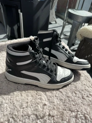 Svarta och vita höga sneakers - Snygga high top sneakers från Puma i svart och vitt. Skorna har klassisk siluett med perforerad tåbox, vita detaljer och Pumas ikoniska formstrip på sidan. Tillverkade i ett mjukt och vädertåligt material med svart snörning och vadderad krage för extra komfort! Så snygga och trendiga, timeless design⭐️Använda men i bra skick utan stora defekter som hål🙌Storlek 39 EU.