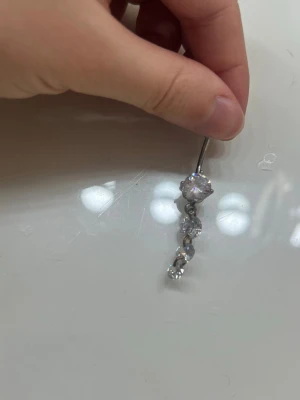 Silvrigt navelpiercingsmycke med stenar - Säljer ett silvrigt navelpiercingsmycke med tre gnistrande, runda stenar i olika storlekar. Staven är i metall och har en kula i ena änden. Perfekt för dig som vill ha något glittrigt och stilrent till din piercing.