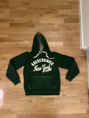Grön Abercrombie hoodie  - Mörkgrön hoodie från Abercrombie med stor vit text 'Abercrombie New York' på bröstet. Hoodien har en stor huva med grått innerfoder och vita dragsnören samt en klassisk magficka. Materialet är mjukt och skönt, perfekt för chill dagar. Det är storlek Medium men den passar också small