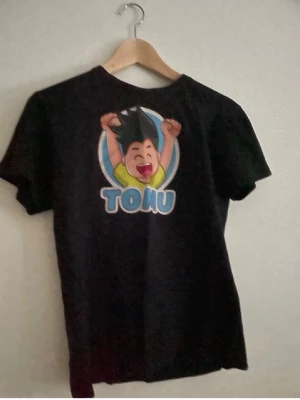 Svart t-shirt med tryck från Clique - Svart t-shirt från Clique med färgglatt tryck på ryggen som visar en tecknad figur och texten 'TONU'. Klassisk passform och rund hals. Perfekt för dig som gillar grafiska motiv och vill sticka ut lite extra.