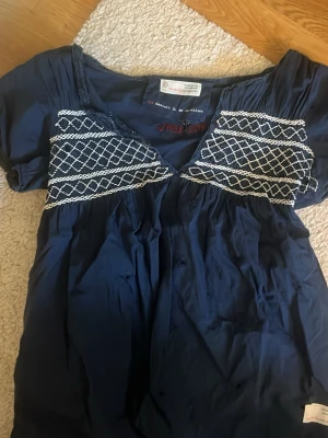 Marinblå kortklänning Odd Molly - Söt marinblå kortklänning från Odd Molly med vita broderade detaljer över bysten och v-ringad hals. Klänningen har korta ärmar och avslappnad passform. Perfekt för dig som gillar bohemiska vibbar och snygga detaljer.