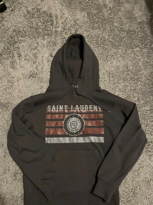 Mörkgrå Saint Laurent hoodie - Snygg mörkgrå hoodie från Saint Laurent med tryck framtill i vitt och rött samt ett emblem i mitten. Klassisk känguruficka och justerbar huva med snören. Perfekt för dig som vill ha en stilren och trendig look. Tillverkad i mjukt bomullsmaterial.