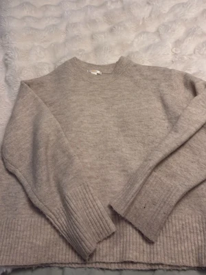 Beige stickad tröja från Gina Tricot - Mysig beige stickad tröja från Gina Tricot med rund halsringning och breda ribbade muddar vid ärmslut och nederkant. Perfekt för kalla dagar och enkel att matcha med olika outfits. Materialet känns mjukt och skönt mot huden.