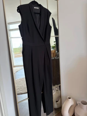 Svart ärmlös jumpsuit med slag - Snygg svart ärmlös jumpsuit med slag i kragen och markerad midja. Byxdelen är rak och har pressveck framtill för en clean look. Perfekt för dig som vill ha en stilren och trendig outfit. Materialet ser ut att vara ett mjukt tyg med lite struktur.
