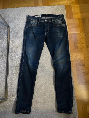 Mörkblå slim fit jeans från Replay - Snygga mörkblå slim fit jeans från Replay med klassiska fem fickor och kontrastsömmar. Jeansen har en lätt tvättad look med slitningar på låren och bakfickor med dekorativa sömmar. Perfekta för en trendig och avslappnad stil.